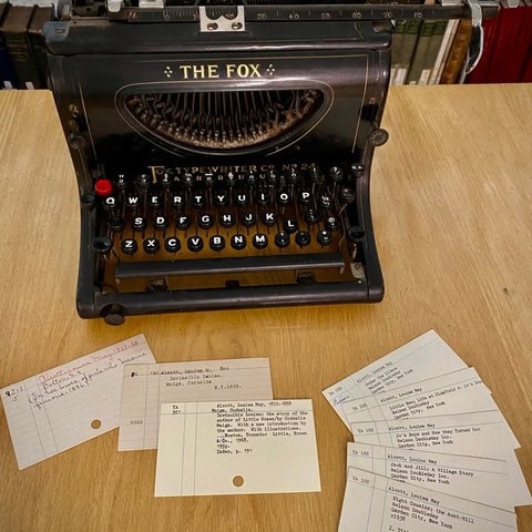an antique typewriter labeled “the fox” sits on a wooden table, surrounded by several typed and handwritten index cards.