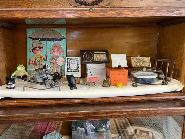 a wooden display case containing vintage collectibles, including a calendar, scale, advertisements, small figurines, and a postcard with illustrated children holding umbrellas.