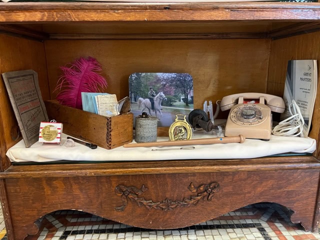 a wooden display case with vintage items including a rotary phone, a feather, old letters, a metal canister, a horse photo, a booklet, and a small trophy.