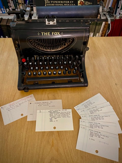 a vintage black typewriter labeled "the fox" is placed on a wooden table, with six library catalog cards spread out in front of it.