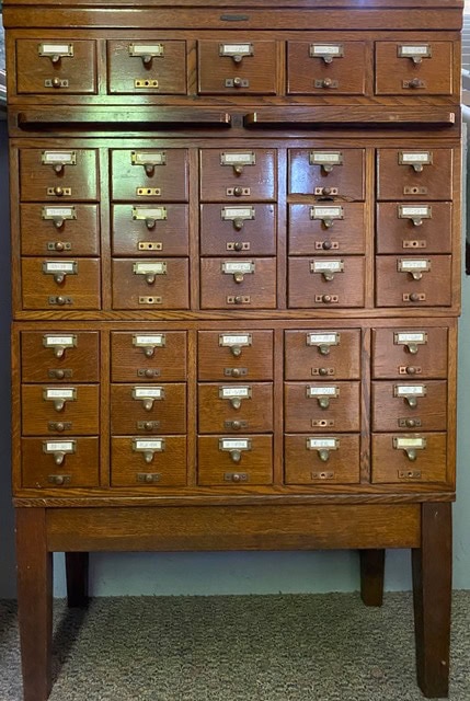 a wooden card catalog cabinet with multiple small labeled drawers, used for organizing and storing index cards.