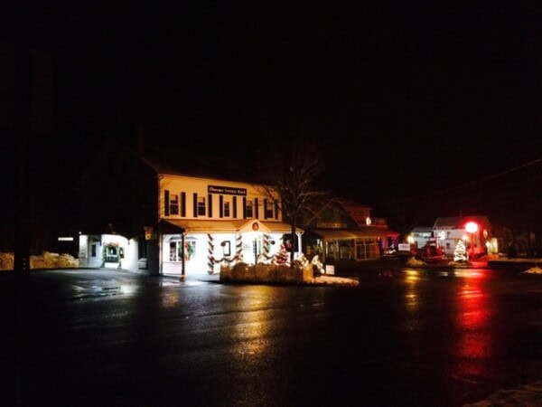a small town street at night with a few buildings and shop windows illuminated, surrounded by darkness and wet pavement.