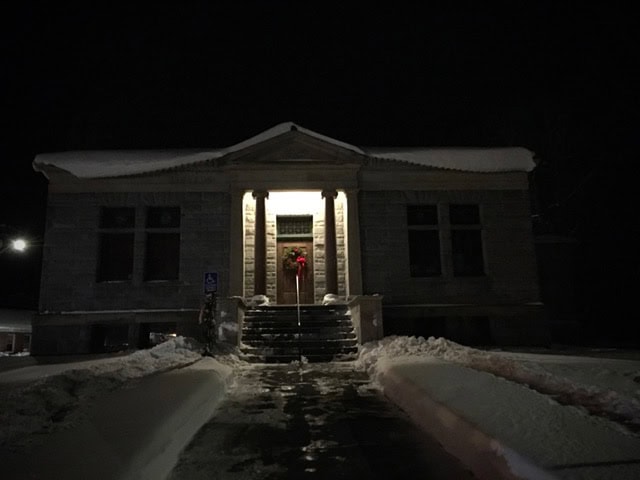 a small stone building with columns and a lit entrance, decorated with a red wreath, is surrounded by snow at night.