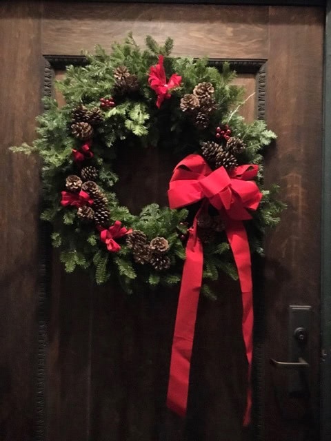 a green holiday wreath decorated with pinecones, red berries, and red ribbons hangs on a dark wooden door near a black door handle.