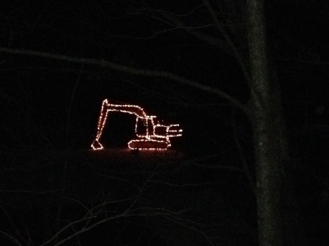 orange string lights arranged in the shape of an excavator or construction vehicle are illuminated in a dark outdoor setting, with tree branches visible in the foreground.