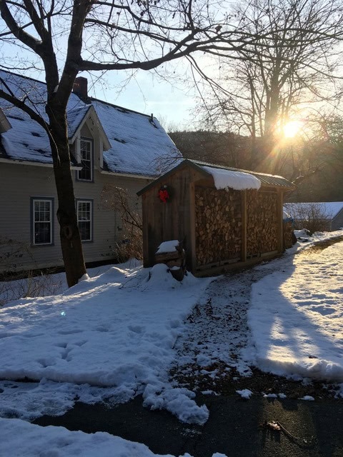 a wood shed stacked with firewood sits beside a house on a snowy day, with sunlight shining through trees in the background.