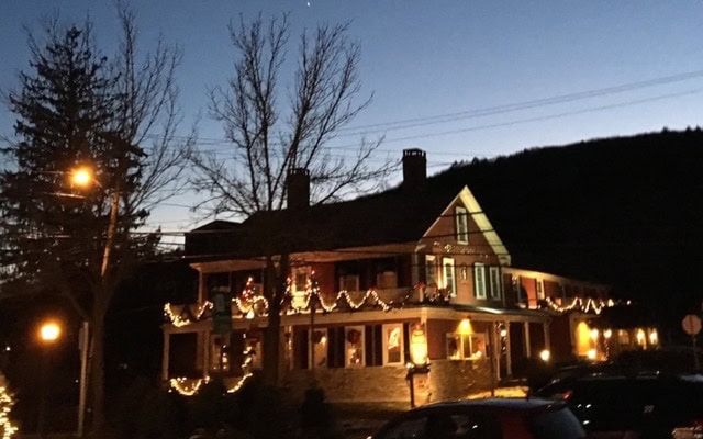 a house decorated with string lights and ornaments for the holidays at dusk, with bare trees and cars parked outside.