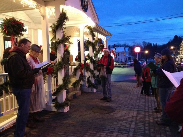 a group of people dressed in winter clothing sing carols outside a decorated building with garlands and wreaths during the evening.