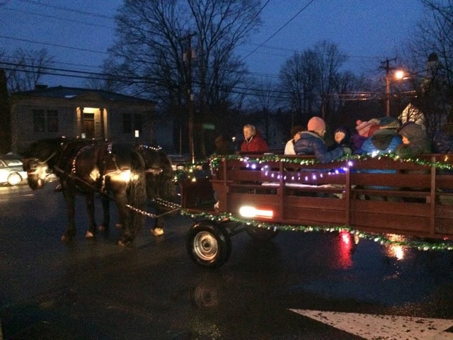 a horse drawn wagon decorated with string lights carries bundled up passengers on a street at dusk.