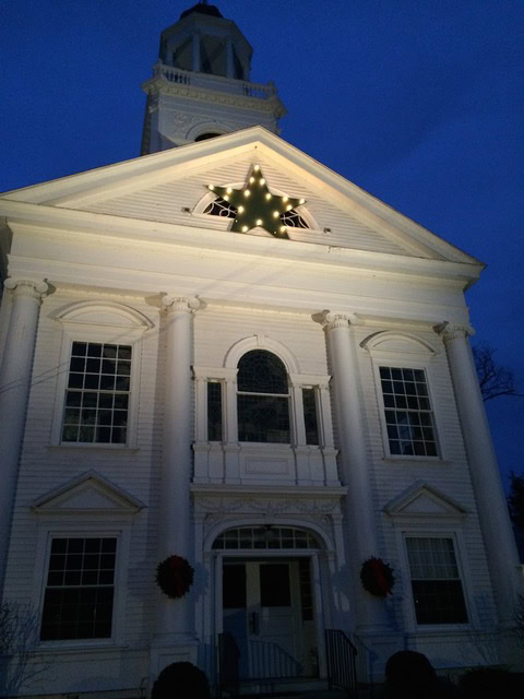 white church building at dusk with a large illuminated star decoration above the entrance and two wreaths on the columns.