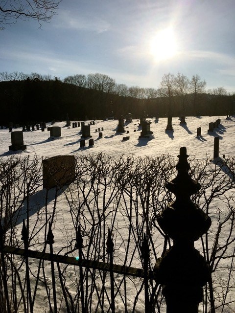 a snow covered cemetery with headstones and leafless trees under a bright sun, viewed through a wrought iron fence.