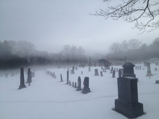 a snowy cemetery with gravestones scattered across the ground, surrounded by leafless trees and shrouded in light fog.