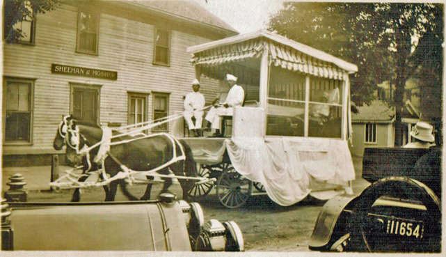 a horse drawn carriage decorated with fabric is driven by two men in white uniforms, passing by a building with a sign reading "sheehan & mosher." vintage cars are in the foreground.