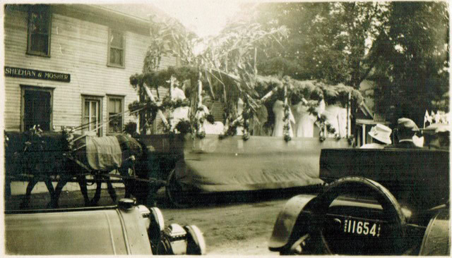 a horse drawn parade float decorated with greenery and flowers passes by a building labeled "sheehan & mosher" as people watch from nearby vehicles.
