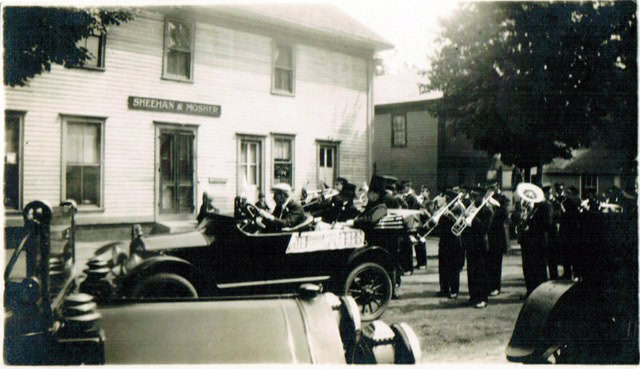 a group of musicians play brass instruments beside an old car outside a two story building labeled "sheehan & monihan.