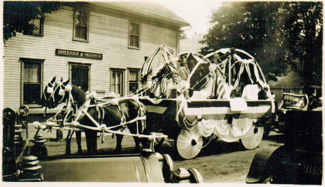 a horse drawn parade float decorated with bunting and streamers passes by a building labeled "sheehan & mosher" in a vintage street scene.