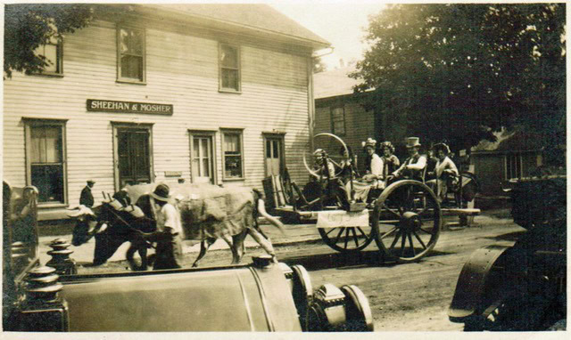 a sepia toned photo shows people in a horse drawn wagon on a street in front of a building labeled "sheehan & mosher," with pedestrians and a car visible.