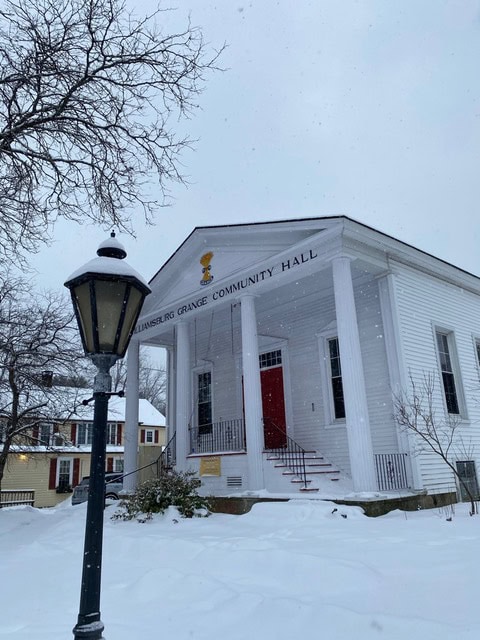 a white, columned building labeled "williamsburg grange community hall" sits in a snowy landscape, with a streetlamp and leafless trees nearby.