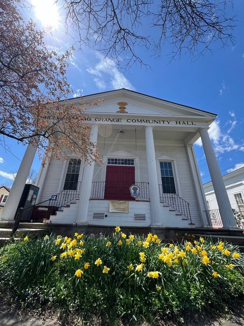white community hall building with four columns, red double doors, and a sign reading "grange community hall." blooming yellow daffodils and a tree are in front. blue sky in the background.