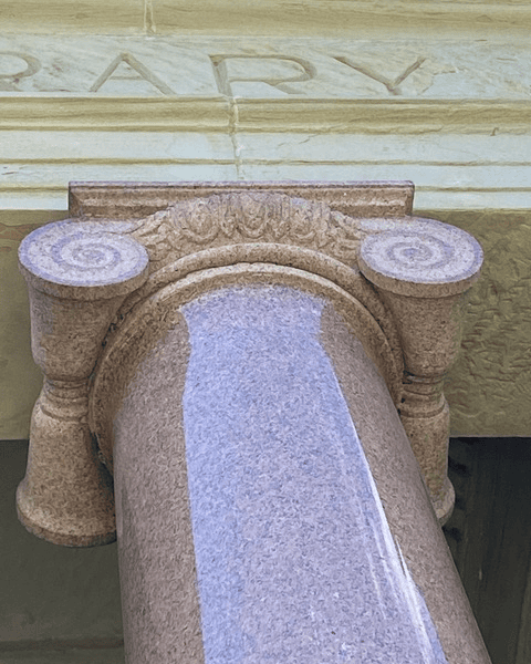 close up of the top of a polished pink granite column with a decorative capital, supporting a stone ledge with partially visible engraved letters.