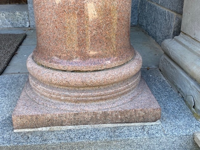 base of a pink granite column on a gray stone step, adjacent to a textured wall and part of a concrete door frame.
