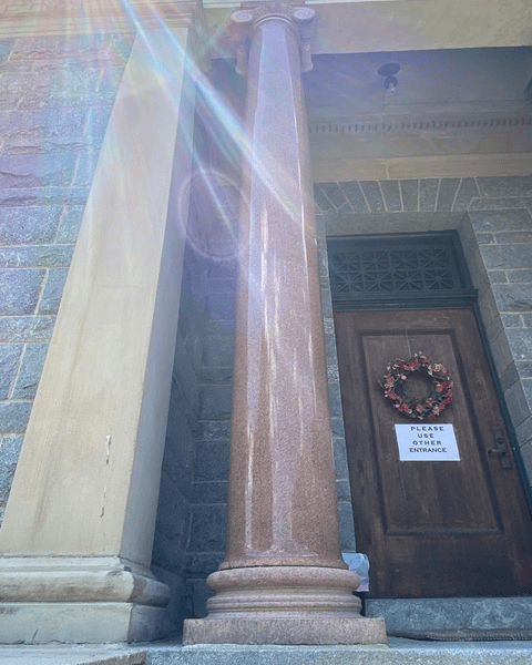 a stone column stands in front of a wooden door with a wreath and a sign that reads "please use other entrance." sunlight creates lens flares in the scene.