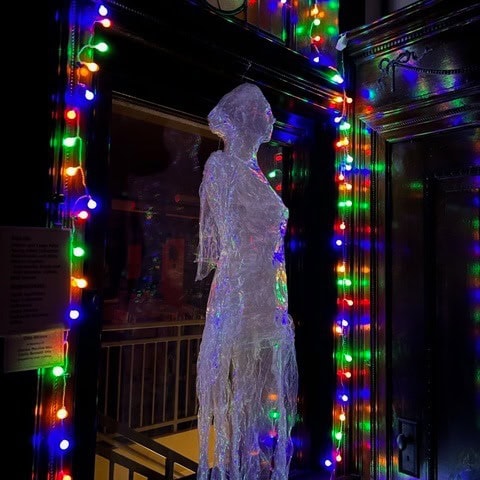 a translucent human shaped figure stands next to a door, framed by colorful string lights in a dimly lit setting.