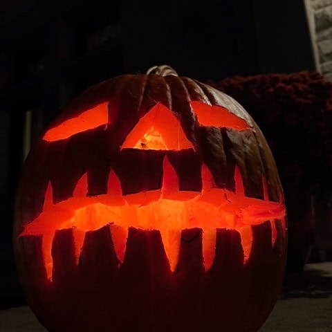 a carved pumpkin with jagged teeth and slanted eyes glows with an orange light in a dark setting.