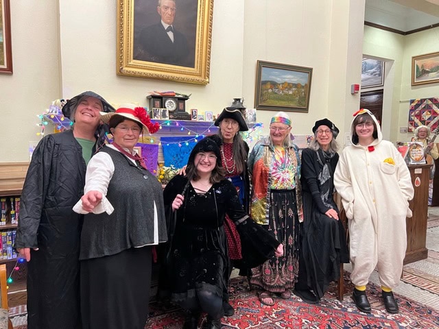 seven adults in various costumes, including witches, a tie dye shirt, and an animal onesie, stand together in a library setting under a portrait and festive lights.