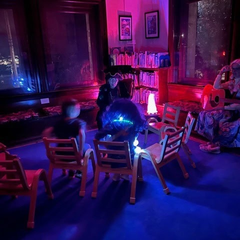 a dimly lit room with purple and blue lighting, several children sit in small chairs, an adult plays guitar, and another adult in a hat interacts with the group near bookshelves.
