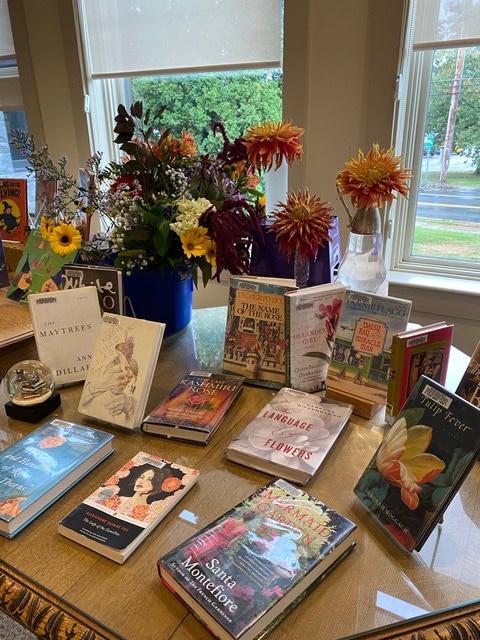 a display of various books is arranged on a wooden table near a window, with bouquets of flowers in vases behind the books.