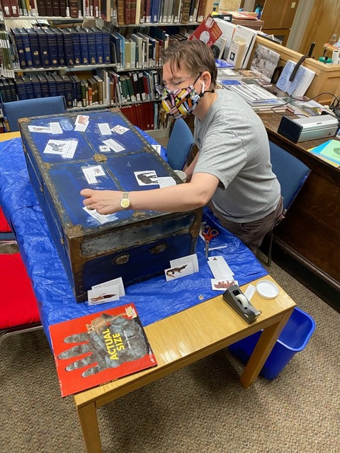 a person wearing a mask works on a large, old blue trunk covered with labels, in a library setting with bookshelves in the background.