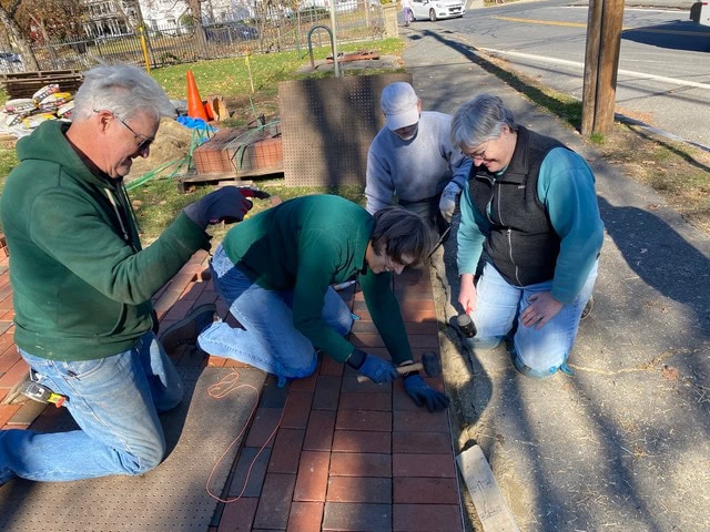 four people work together laying bricks on a sidewalk, using tools to position and secure the bricks in place on a sunny day.