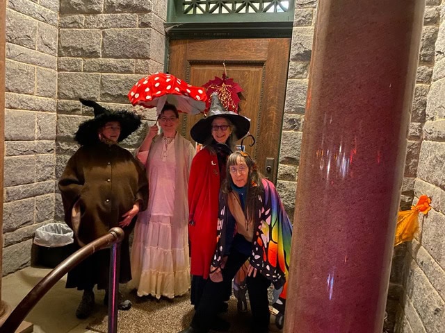 four people in costumes stand outside a stone building; one wears a black cloak, one a white dress, one a red cape with an oversized mushroom hat, and one a colorful shawl.