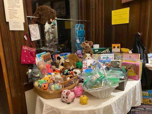 a round table displays assorted plush toys, board games, children's books, and small gifts in a wood paneled room, with a stuffed mammoth hanging above.