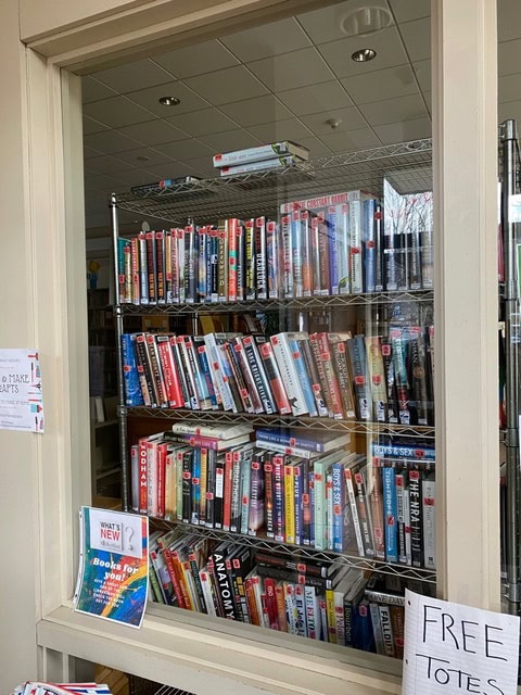 a metal bookshelf filled with assorted books is visible through a window. a "free totes" sign and a "what's new?" display with books are positioned outside the window.