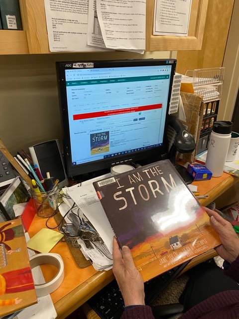 a person holds a book titled "i am the storm" in front of a cluttered desk with a computer displaying a library catalog entry for the same book.