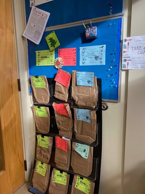a display rack holds brown paper bags labeled with colorful sheets, next to a bulletin board with flyers and a craft kit notice.