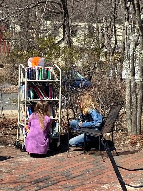 two girls are selecting books from a rolling book cart outdoors on a sunny day, with one sitting on a metal chair and the other crouching by the cart.