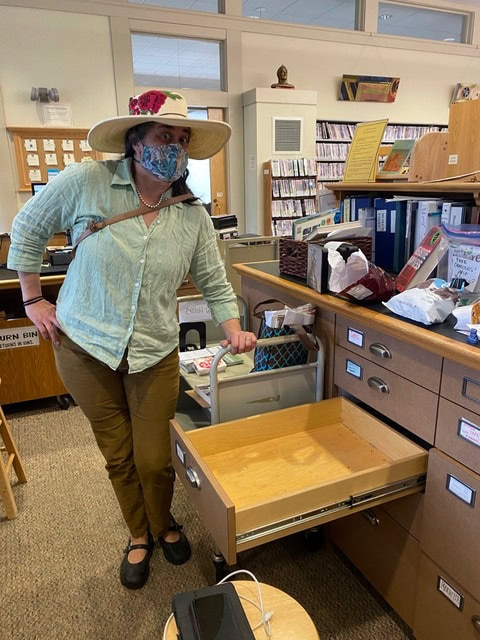 a person wearing a hat and face mask stands next to an open drawer in an office or library setting.