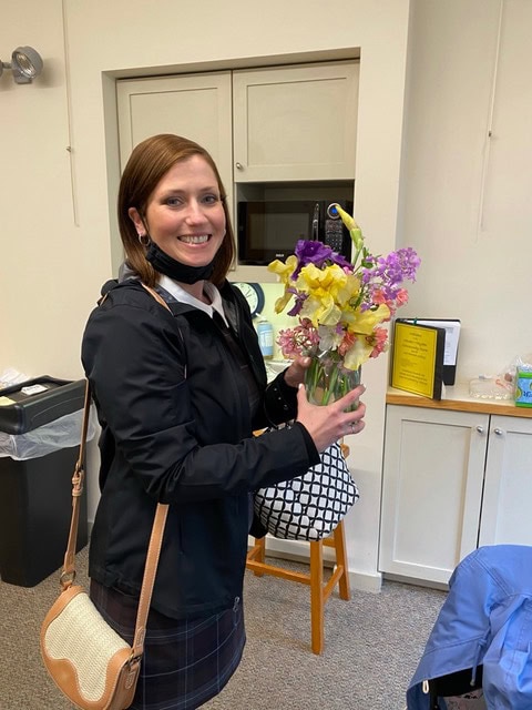 a woman standing indoors holds a vase of colorful flowers and smiles at the camera. she wears a black jacket and has a white bag over her shoulder.