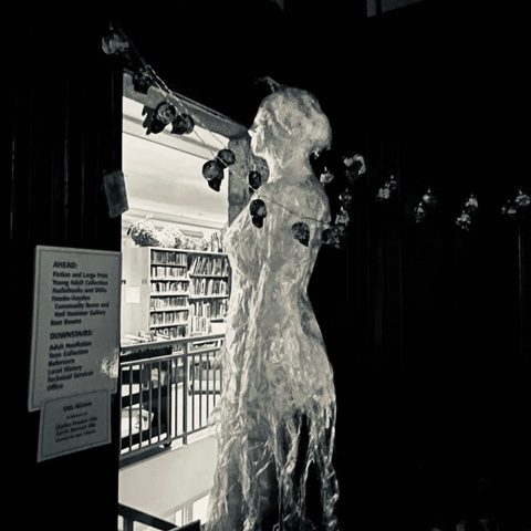 a translucent sculpture of a human figure stands near a doorway decorated with a string of small skulls, with bookshelves visible in the background.