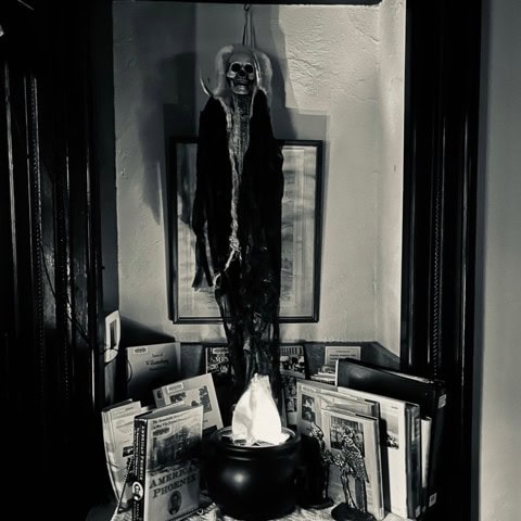 a tall skeleton decoration hangs above a cauldron surrounded by books and newspapers in a dimly lit corner.