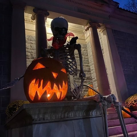 a large skeleton sits on stone steps behind a glowing, carved jack o’ lantern, with columns and a stone building in the background.