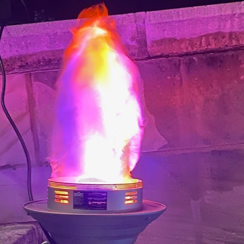 a portable heater emits a tall, colorful flame that transitions from orange at the top to purple and white at the base, set against a stone wall at night.