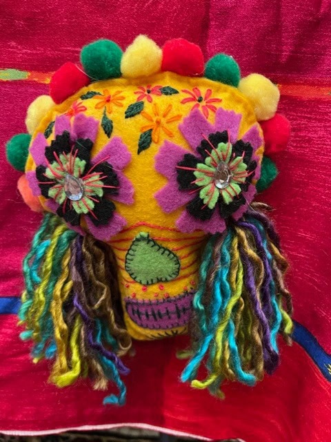a colorful felt skull decorated with yarn hair, pom poms, and fabric flowers, set against a bright pink background.