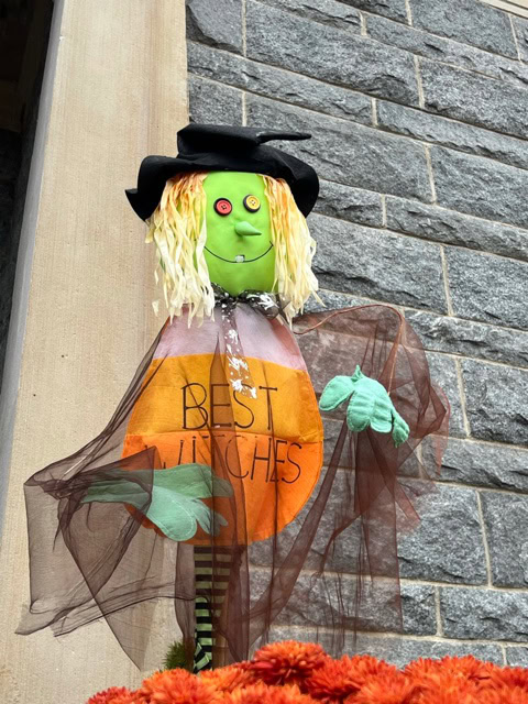 a green faced witch decoration with yellow hair and a black hat stands against a stone wall, wearing a sign that reads "best witches." orange flowers are visible at the bottom.