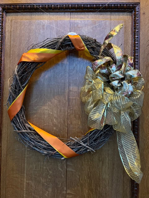 a grapevine wreath with an orange ribbon and a large gold decorative bow hangs on a wooden door.