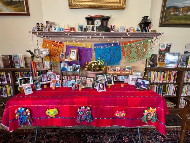 a day of the dead altar with framed photos, candles, flowers, papel picado, and decorations displayed on a table and mantel in a room with bookshelves and paintings.