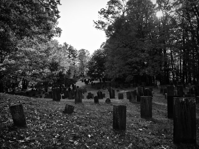 black and white photo of an old cemetery with numerous weathered, upright gravestones on a grassy slope surrounded by trees.
